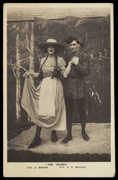 Download the full-sized image of Two members of a military concert party pose on stage: one is in drag with a large hat and pigtails. Photographic postcard, 1915-1916.