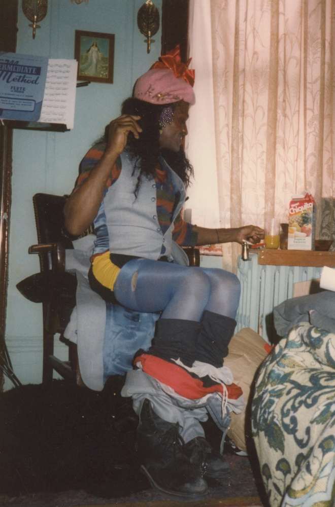 Download the full-sized image of A Photograph of Marsha P. Johnson Sitting in a Chair Wearing a Pink Hat, Blue Vest, and Blue Tights